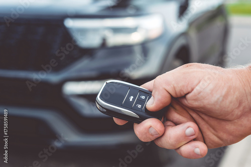 Close-up of a man unlocking a car using a key fob, with a black car parked in a car park in the background.
