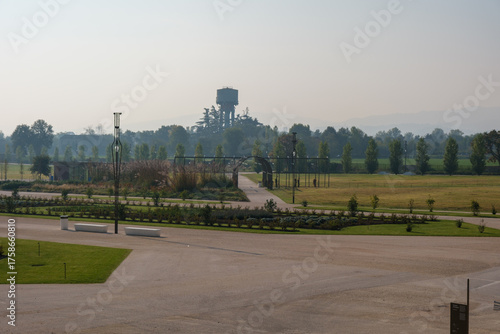 A wide gravel promenade in the Reggia di Rivalta garden, lined with rows of trees casting long shadows, leads to a distant crowd enjoying the spacious park grounds on a bright day. High quality photo