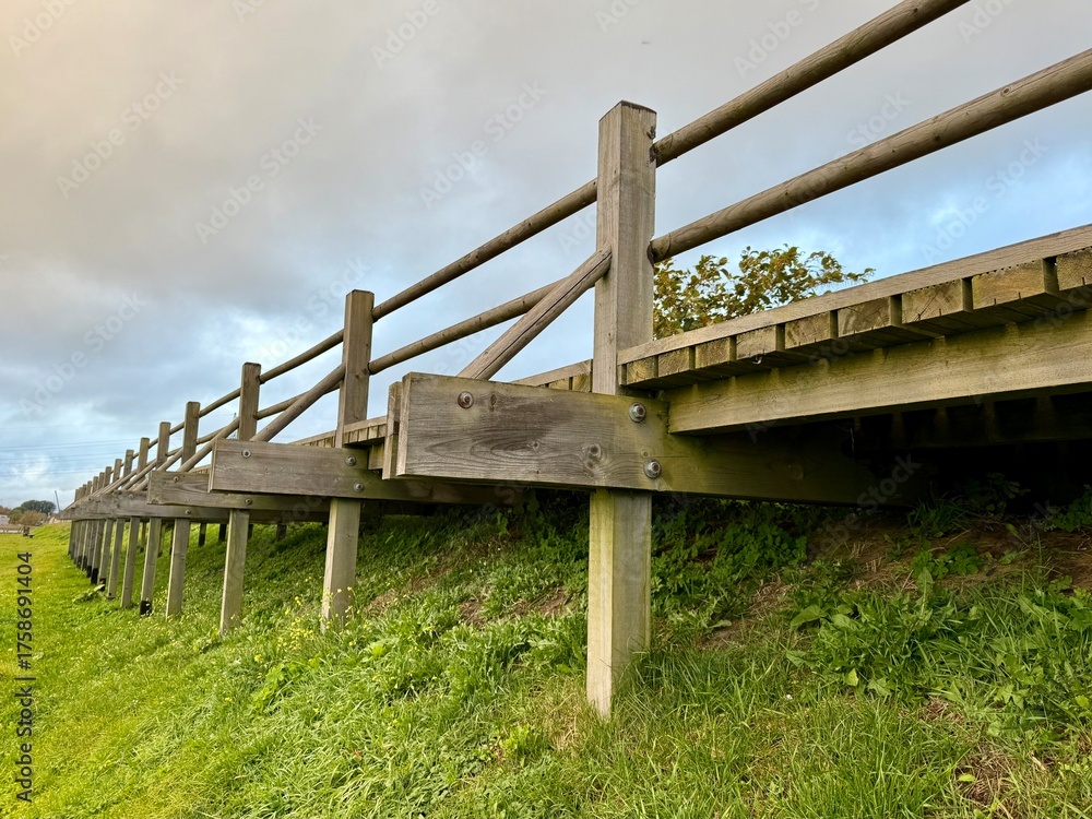 Fototapeta premium Low-angle view of a wooden footbridge with railings standing on grassy ground under a cloudy sky.