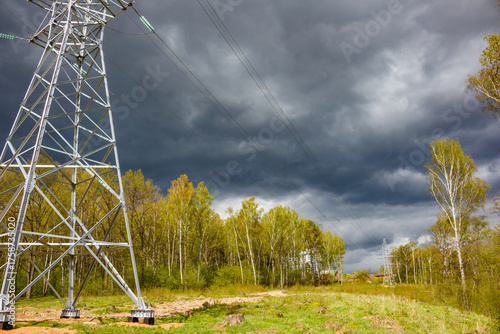 Sturdy electricity pylon commands the foreground, silhouetted against a brooding, stormy sky. Vibrant green spring foliage lines the horizon