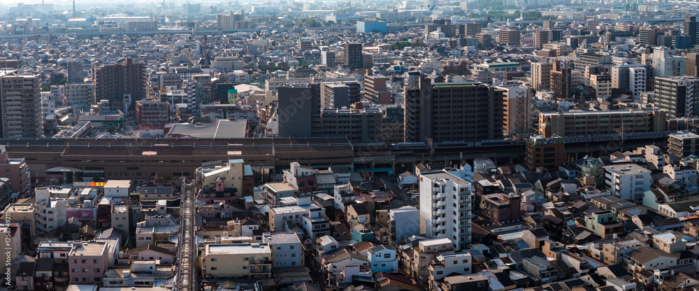 Naklejka premium Aerial view of Osaka, Japan, shows mid rise apartments, tight houses, and an elevated railway with a passing train in hazy afternoon light across a vast district.