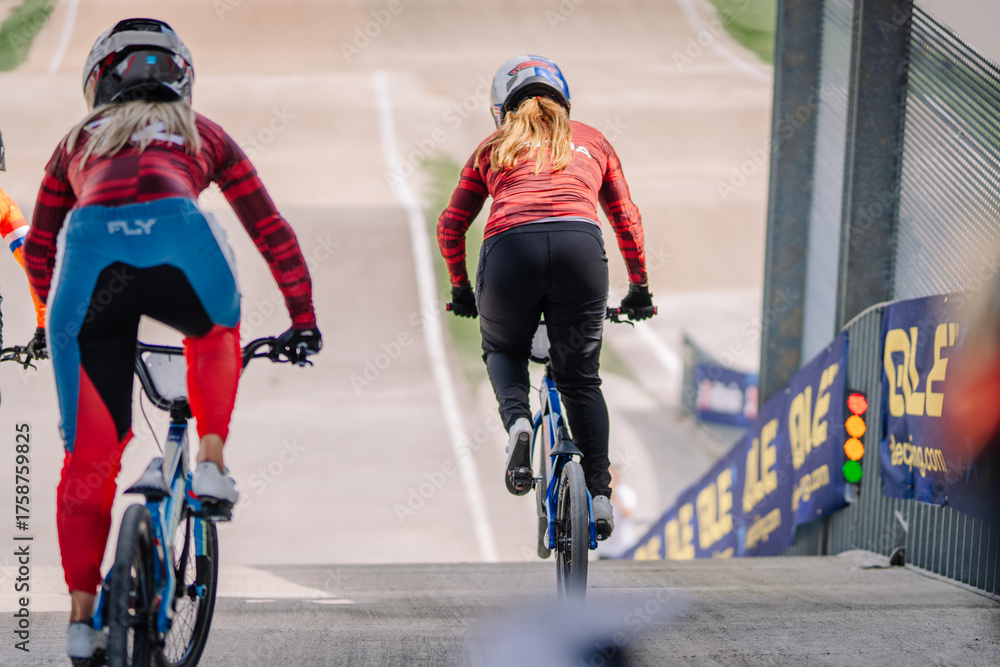Fototapeta premium Valmiera, Latvia - July 12, 2025: Female BMX riders in colorful jerseys preparing to race on a professional track with vibrant surroundings