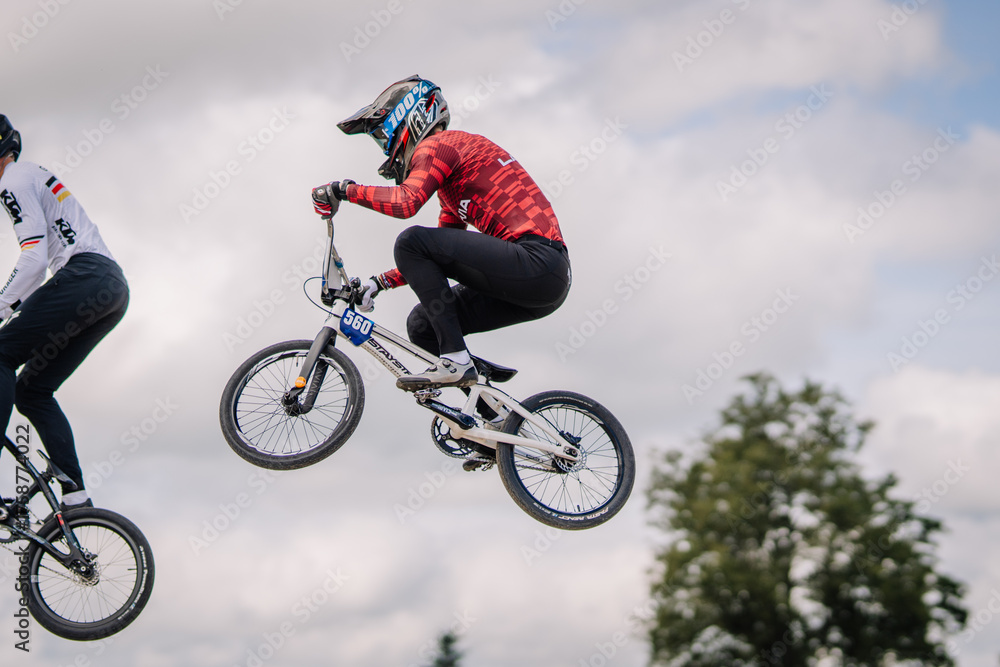 Fototapeta premium Valmiera, Latvia - July 12, 2025: Young male BMX rider in red jersey performing a jump on a bicycle during a competition, showcasing athleticism and excitement in a dynamic outdoor setting