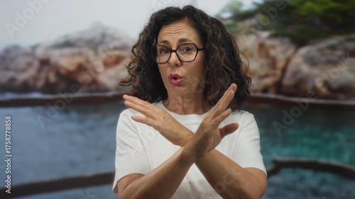 Middle age hispanic woman crosses arms in rejection gesture on rocky beach; denial refusal stubbornness.