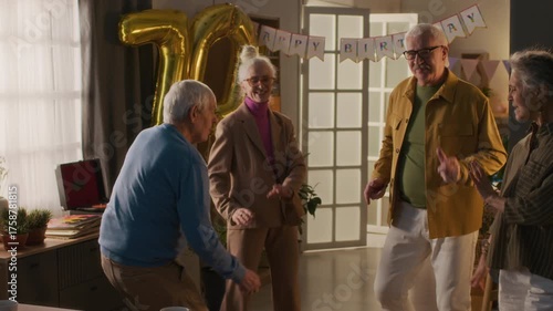 Company of four cheerful senior friends energetically dancing during birthday party of 70-year-old lady wearing pantsuit