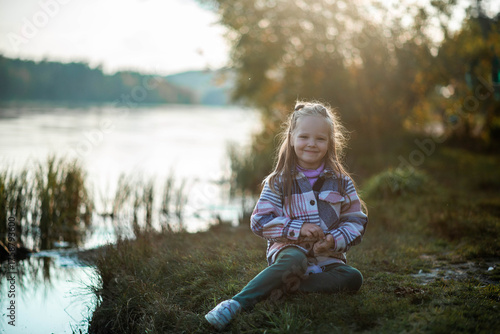 Little girl sitting by the lake and looking at the water during autumn evening. Peaceful moment in nature with warm sunset light, calm reflection, and serene countryside atmosphere.