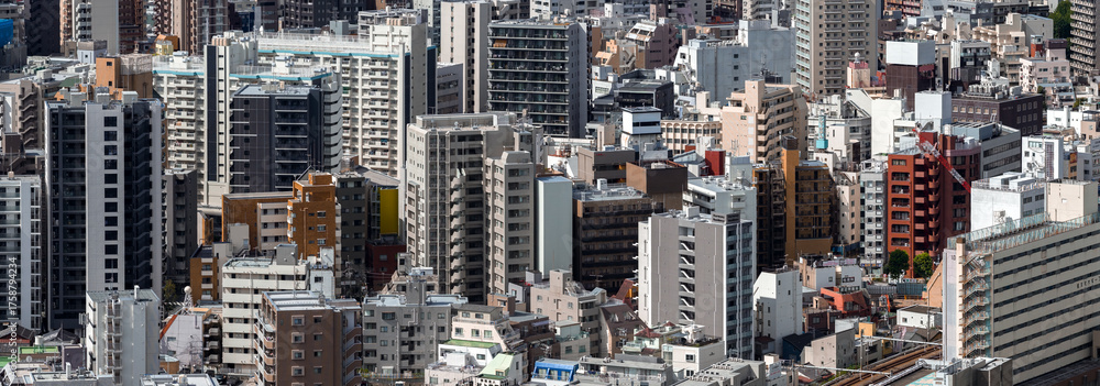 Obraz premium Central Tokyo view shows dense mid and high rise buildings, varied modern facades, rooftop gear, balconies, and a crane, with rail tracks visible at lower right in daylight.