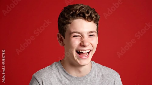 A young individual with curly hair laughs with eyes closed against a vibrant red backdrop