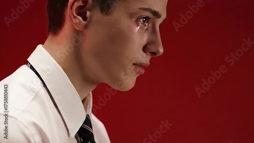A young man cries, his head bowed, wearing a white shirt and a black tie, against red