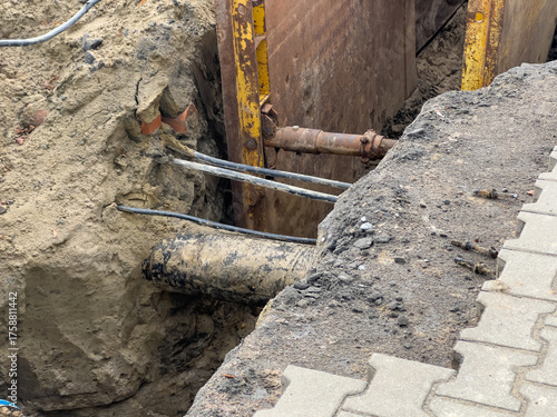 Open construction trench exposing utility pipes and electrical cables, secured by metal shoring for urban infrastructure repair and maintenance
