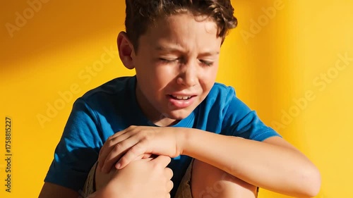 A young boy, expressing pain, eyes shut, hands on knees, against a vibrant yellow backdrop