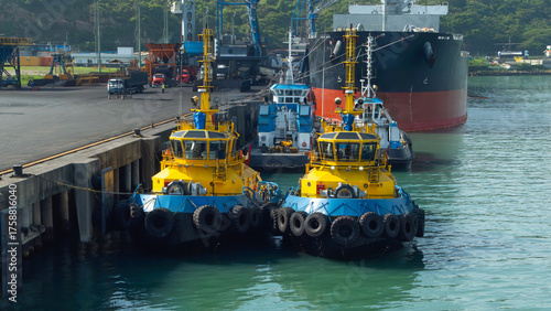 Bright yellow tugboats moored at a busy port alongside a large cargo ship. Industrial maritime scene showcasing harbor operations & logistics