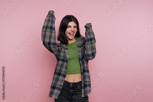Young woman celebrating joyfully against a vibrant pink background while wearing a casual outfit with a plaid shirt