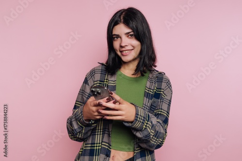 Young woman smiling while holding smartphone in front of pink background during casual indoor setting