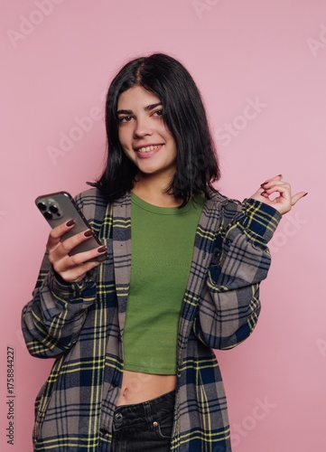 Young woman in green top and plaid shirt smiles while using phone against pink background