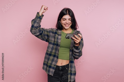 Young woman celebrates success while looking at her smartphone against a pink background
