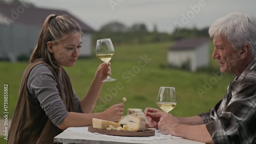 Side view shot of young Caucasian female farmer sitting at table with her senior father outdoors and discussing flavor of artisan cheese varieties made in their creamery