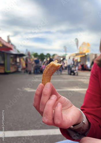 Closeup of hand holding a churro snack at an amusement park with blurred background of colorful food stands and people walking.