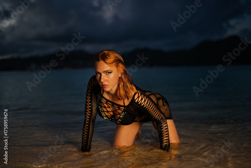 
Woman in a black fishnet dress over a bikini standing in the ocean at dusk, with mountains and a stormy sky in the background.