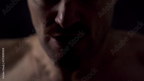 Close-up portrait of an athletic man against a dark background. He hits himself on the chest with his fist several times.