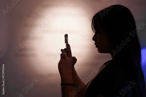 Silhouette of a black-haired young girl holding a smartphone in her hand, looking forward into the room.