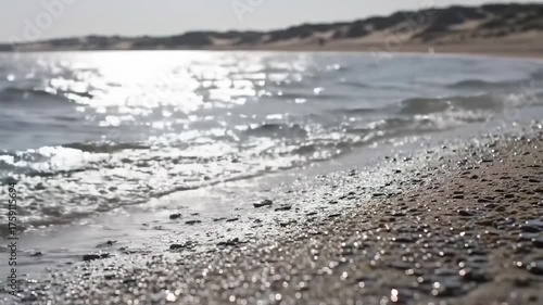 Sunlit water laps a grainy shore, blurring to a sandy bank and distant dunes