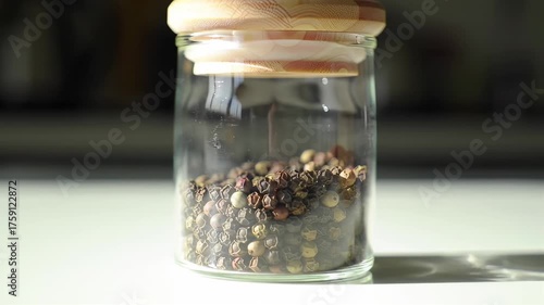 A clear glass jar filled with peppercorns, topped with a light wood lid, on a white surface