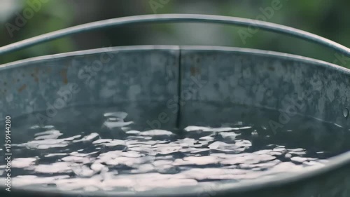 A close-up shot of a metallic bucket partially filled with rippled water against blurred greenery