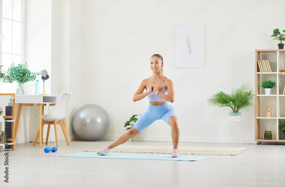 Fototapeta premium Happy beautiful fit young Asian woman in yoga shorts doing a side lunge exercise on a workout mat in a living room interior at home. Sport, fitness, healthy lifestyle concept