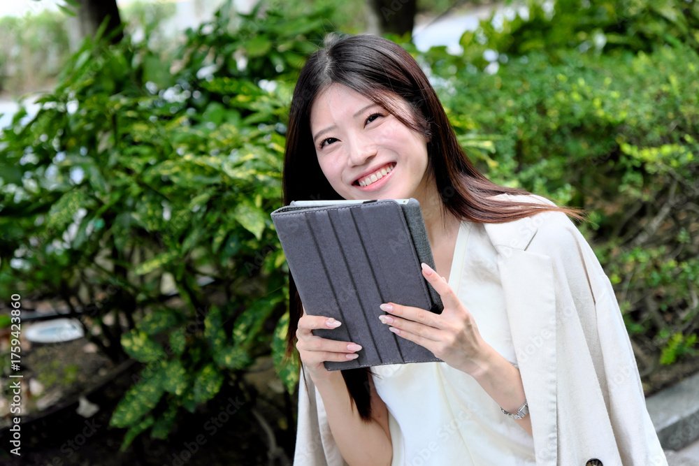 Fototapeta premium 屋外の緑に囲まれ笑顔を見せる、タブレットPCを持つアジア人女性のポートレート。リモートワーク、デジタルライフ、ビジネスとリラックス、テクノロジー、環境調和を表現するイメージ素材、フレッシュな雰囲気で採用広告にも最適