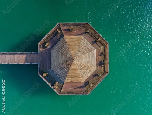 Aerial view of a wooden octagonal pavilion extending over vibrant turquoise water, connected by a narrow walkway.