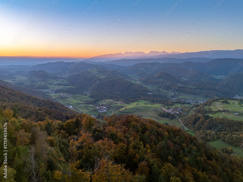 Naklejka premium Panoramic view over the Zasavje region’s rolling hills and valleys at dusk, with an autumn forest in the foreground and the distant Kamnik-Savinja Alps silhouetted on the horizon.