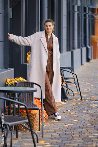 Woman in stylish outfit enjoys autumn day by sidewalk cafe amid colorful fallen leaves