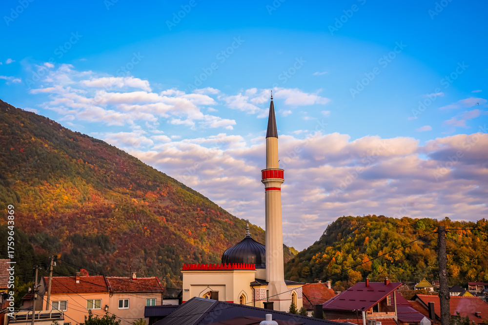 Fototapeta premium Beautiful mosque with tall minaret surrounded by autumn-colored hills and village houses in Travnik, Bosnia and Herzegovina, under a bright blue sky with scattered clouds.