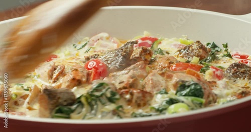 Chef cooks tomatoes, spinach, and salmon in creamy sauce, stirring with a wooden spatula. 4K close-up of home cooking process.