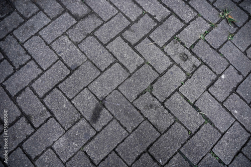 Top-down view of grey paving stones in a herringbone pattern. Pavement block texture background with weeds