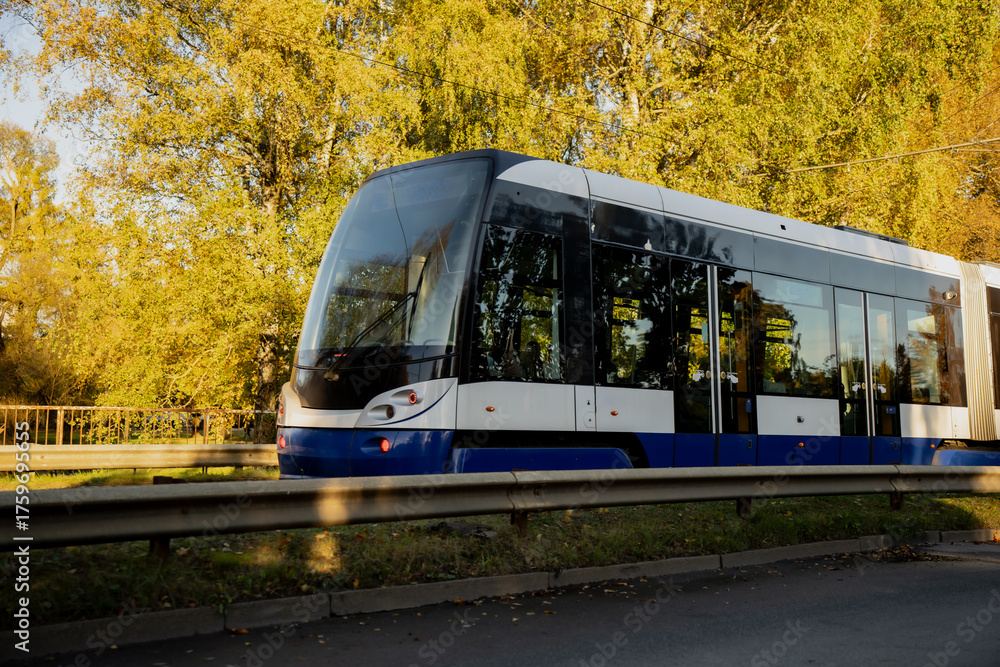 Fototapeta premium Modern blue and white electric tram moving along city track surrounded by autumn trees in Riga, showing contemporary public transportation design and sustainable urban mobility