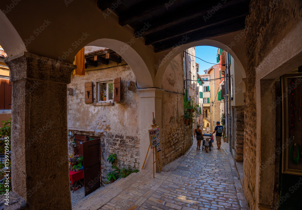 Fototapeta premium Rovinj, Croatia - August 14, 2025: Arched passageway opens to a sunlit cobblestone street lined with rustic walls and wooden shutters, where a small family strolls through the quiet charm