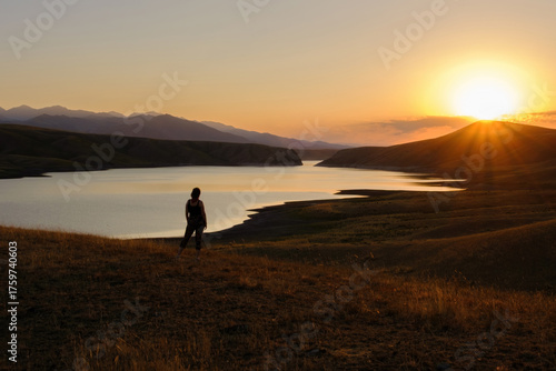Canvas Print Silhouette of a woman on a hill enjoying the picturesque view of the mountains and water at sunset