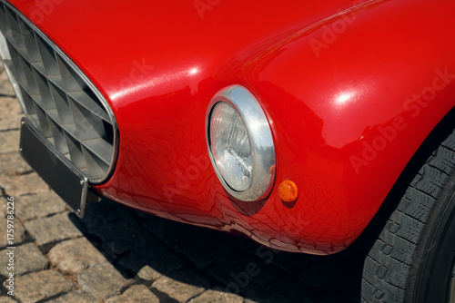 Close-up of a vintage red sports car's front fender, showing the chrome headlight and patterned grill on a cobbled street.