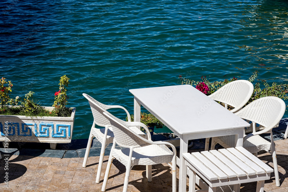 Fototapeta premium A Tranquil Waterfront Scene Featuring White Furniture and Calm Waters Surrounded by Lush Greenery and Colorful Flowers
