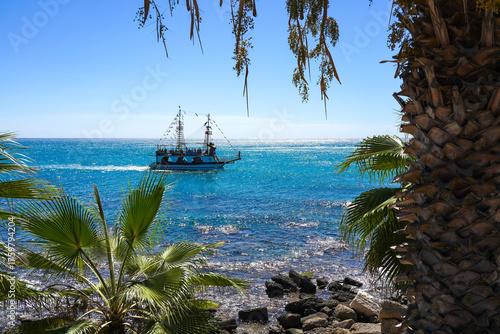 A picturesque view of a sailing ship navigating through vibrant turquoise waters, framed by lush palm trees under a clear blue sky on a sunny day.