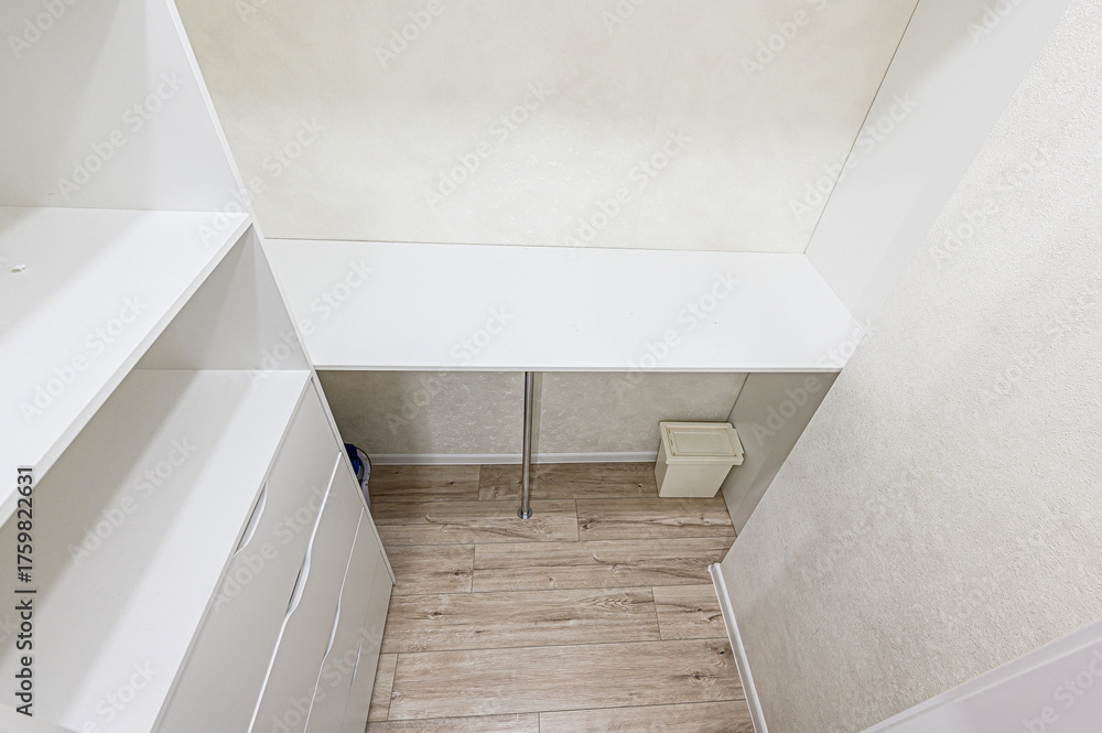 Fototapeta premium A closet space with white shelving, drawers, a desk area, and a trash bin on a wood-look floor