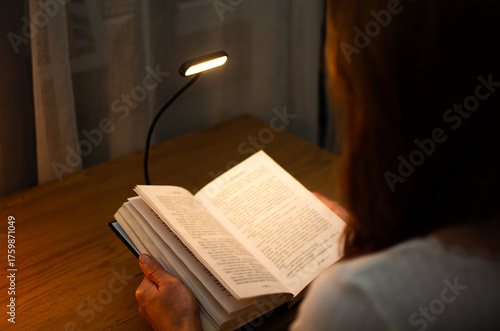 A woman is reading a book at a wooden table in the evening with the help of convenient clip-on lamp