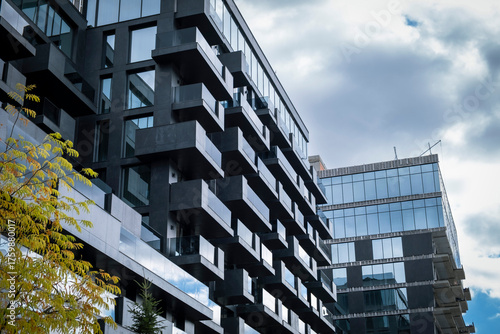 Fotografie Modern, dark gray facade of a high-rise residential building with projecting balconies and large glass windows, set against a dramatic, cloudy sky, with a touch of autumn foliage