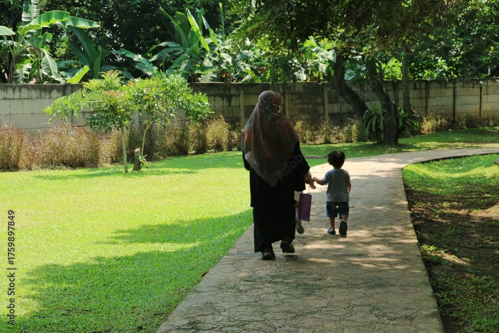 Fototapeta premium Mother Wearing Abaya Walks Hand in Hand With Child Along Park Path Under Green Trees