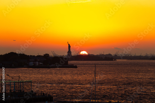 Sunset over New York Harbor with the Statue of Liberty silhouetted against the vivid orange and yellow sky with an airplane flying in the distance