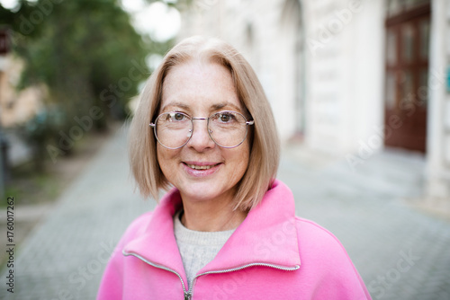 Smiling middle aged woman 50-55 year old wearing eye glasses and pink sweatshirt walking in city street outdoors over sun light. Happiness.