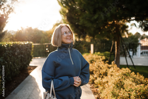 Smiling senior woman 55-60 year old wearing eye glasses walking in city street outdoors over sun light. Happiness.