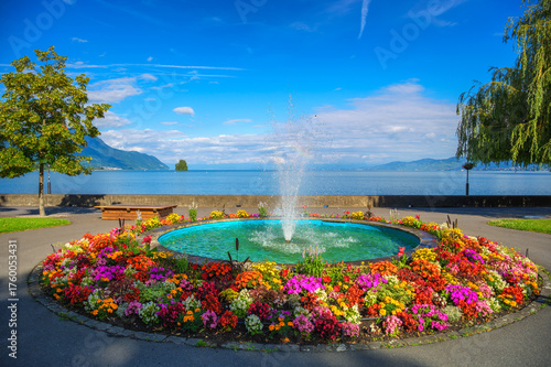 Circular fountain surrounded by vibrant flowers with Lake Geneva and mountains in the background, Villeneuve, Switzerland.